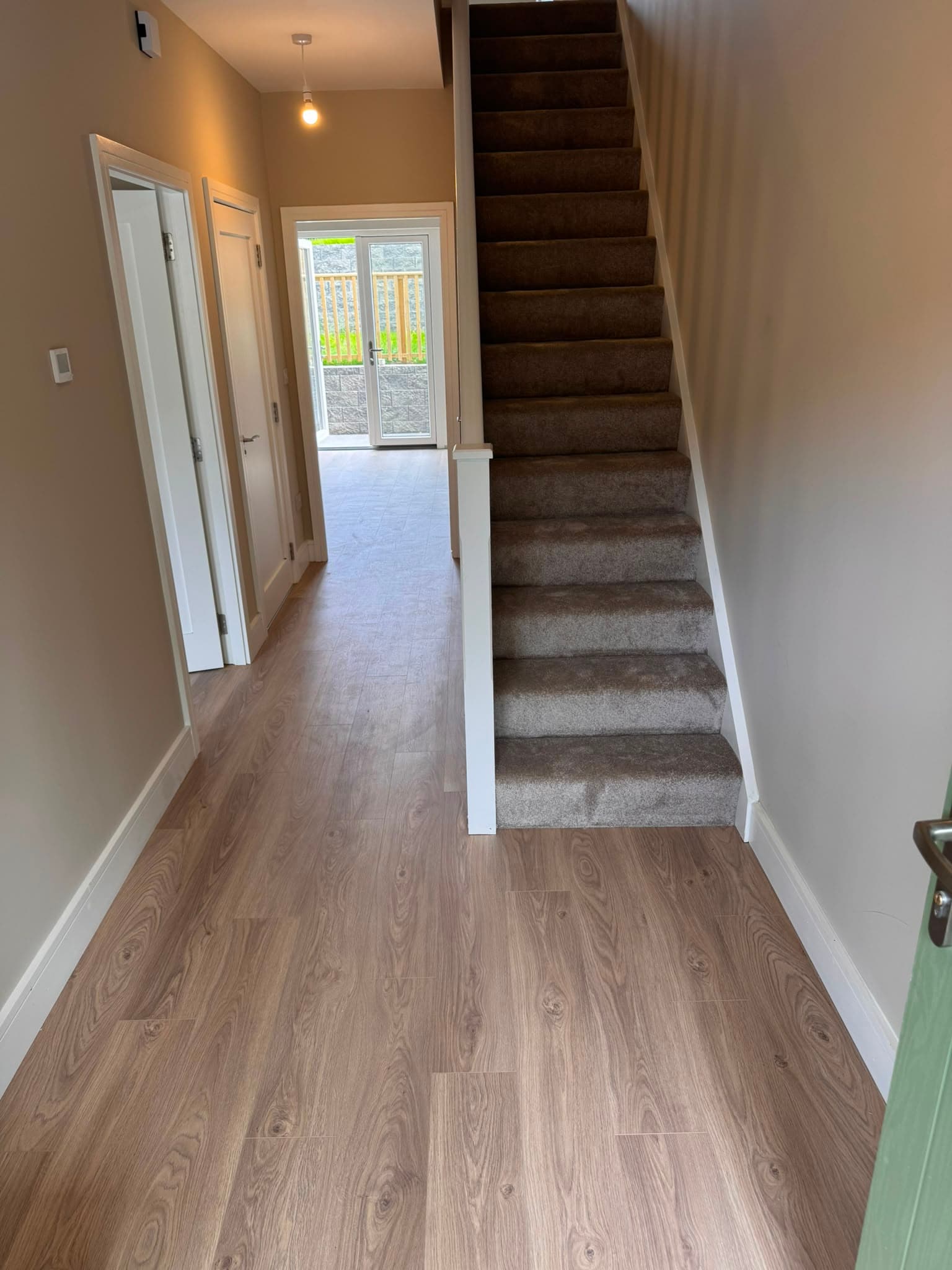Warm-toned laminate flooring fitted through a hallway with a carpeted staircase leading off it